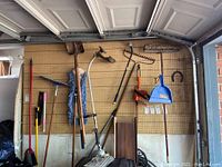 Wide view of garden tools including shovels, rakes, broom, dustpan, hedge trimmer, and weed eater hung on pegboard in garage.