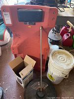 Red 6-wheeled mechanic's trolley with cushioned headrest, with nearby white bucket and long metal rod attachment of driveway cleaner.