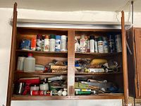 Three wooden shelves filled with assorted spray paints, bottles, sandpaper, rollers, and painting supplies viewed from the front.