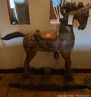 Side view of hand carved wooden rocking horse showing colorful painted saddle, bridle and textured mane. Tail is broken.
