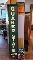 Tall green wooden Quaker State Motor Oil advertising sign leaning against wall and furniture indoors.