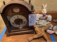 Photo showing Tempus Fugit mantel clock, porcelain bunny doll with floral dress, and small wicker woven figurine on wooden surface with textured wall backdrop.