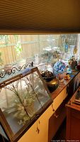 Window sill display showing metal and glass terrarium with decorative scrollwork and hanging charms, ceramic pots with plants, candles, and glass decor.