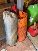 Two rolled tarps, one grey-beige and one bright orange, placed upright on the floor with gas containers in background.