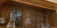 Shelf display of eight Waterford crystal couple figurines, candy dish with lid, decorative vase filled with rocks, and four wine glasses.