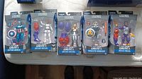 Five Marvel Legends Captain America Series figures displayed in box packaging showing detailed sculpt, colors, and accessories including shields and weapons.