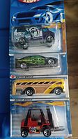 Four Hot Wheels cars in packaging: purple 'SaberTooth' van, black flame car, yellow woody wagon, red forklift truck.