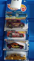 Five unopened Hot Wheels cars in original packaging, different models and colors, stacked vertically in plastic trays.