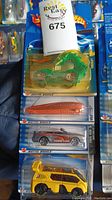 Four Hot Wheels cars in original packaging stacked, showing green robotic car on top, orange and silver cars, and yellow van car at bottom.