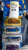 Four unopened Hot Wheels cars in original packaging displayed on a surface. The cars include different models and colors: an orange and gold streamlined car, a blue futuristic car, a purple muscle car, and a Chrysler Pronto with American flag design.