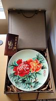 Ceramic plate with floral design in box alongside pamphlet and satin lining