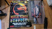 Two posters rolled out on a wooden floor including a Mortal Kombat promotional poster with character portraits and a Yokohama racing poster featuring a race car.