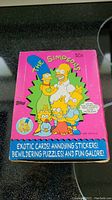 Front view of sealed Simpsons Topps collector card booster box with cartoon image of Simpsons family and text about exotic cards and stickers.
