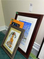 Two framed prints leaning against a wall on the floor. The closest one is a colorful print with a grayish wooden frame titled 'Sugar Spice.' Behind it is a larger framed print of a church in a winter setting with a dark brown wooden frame.