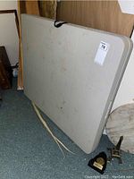 Beige rectangular folding table leaning against wall showing side and top surface, with black carrying handle visible.