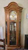 Front view of solid oak grandfather clock with glass panels showing pendulum and weights inside.