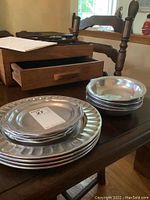 4 pewter dinner plates and 4 salad plates stacked separately on a wooden table with a drawer unit in background.