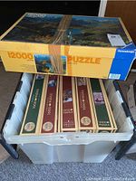 Top view of the set showing the large Ravensburger puzzle box on top of a plastic storage bin containing 5 unopened Cobble Hill 1000 piece puzzles.