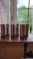 Photo of six cylindrical pewter steins with wooden handles and bases on a wooden table by a window.