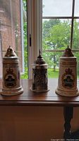 Three beer steins displayed together, two ceramic with military insignia and one highly detailed pewter stein in center