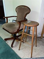 Vintage upholstered swivel office chair in brown patterned fabric with wooden carved arms and a swivel base with caster wheels next to a wooden stool with round seat and four legs.