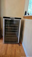 Front view of the Danby wine fridge showing glass door with metal racks inside, placed on a wooden floor against a wall.