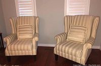 Pair of wingback armchairs upholstered in beige and cream vertical stripes with matching square throw pillows, placed side by side on hardwood floor.