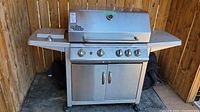 Full view of stainless steel Grill Chef propane BBQ grill with 5 main burners, side burner, and storage cabinet underneath. Grill is on wheels and situated on deck with a wooden fence background.