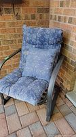 Metal outdoor chair with blue patterned cushioned seat and backrest, matching throw pillow, placed on brick patio against brick wall.