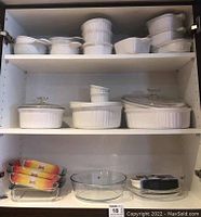 Cabinet shelves with assorted CorningWare French White ceramic bakeware dishes, some with glass lids, ceramic bowls with handles, and storage containers on three shelves.