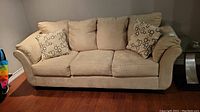 Front view of beige microsuede 3-seat sofa with three seat cushions and matching back cushions, including two decorative floral pattern throw pillows, on a wooden floor.