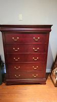 Front view of wood dresser showing five drawers with brass handles, reddish-brown finish, and decorative molding on top and base.