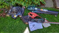 Three electric yard tools placed on grass outdoors: Black & Decker electric mower, orange and black Yardworks hedge trimmer, green and black Yardworks leaf blower. Manuals and papers next to hedge trimmer.