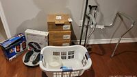 Photo showing foot massager circulation booster, boxed supplies, laundry basket, over toilet aid, cane, and walker against wall on hardwood floor
