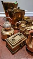 Various brass and copper decor pieces arranged on floor showing variety and details such as buckets, kettle, candle holders, and containers