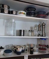 Full view inside kitchen cabinet showing shelf with mugs, storage bowls, seasoning accessories, and glass pitcher.