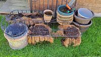 Photo showing assorted planters and garden tools arranged outdoors on grass and concrete. Items include a large metal wire frame planter with coconut coir liners, multiple ceramic and plastic planters, and hand garden tools: a small rake and a trowel.