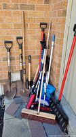 Full view of all garden tools including shovels, brooms, and snippers leaning against a brick wall.