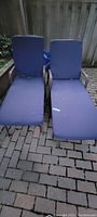 Pair of outdoor lounge chairs with blue cushions on a brick patio in front of a wooden fence.