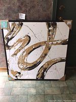 Abstract canvas artwork with swirling black and gold paint on white background, framed in wood, measuring 34.5 x 34.5 inches.