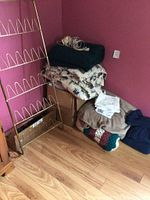 Photo of folded bedding and blankets stacked next to a gold metal shoe rack and a bundle of hangers on a wooden floor against pink walls.