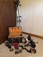 Photo showing multiple lenses, chrome frame filters, camera cases and adjustable tripod standing upright against wall