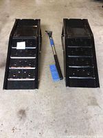 Top-down view showing two metal car ramps placed side by side with a snow brush in the middle, visible condition and details of the ramps and brush.