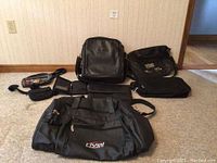 Multiple carry bags of various sizes and styles including a large rivel duffel bag, two square/rectangular work bags, a business card/agenda holder, and smaller black bags laid on floor.