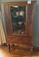 Front view of wooden china cabinet showing glass door with decorative wooden grid and carved panels on sides plus carved drawer beneath.