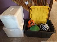 Two white styrofoam coolers stacked with large black tote with yellow lid containing small lunch coolers and hats with white wire hat dryer