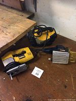 DeWalt jigsaw with yellow body, black cord, and saw blade lying on wood surface with black and yellow hard case lantern behind it
