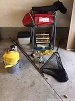 Overall lot arranged showing 2 fishing rods, fishing net, tackle box with lures, 2 folding chairs, small cooler, bucket, and yellow minnow cage.
