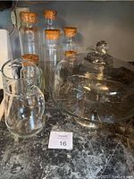Glassware set with seven glass containers with corks, a glass pitcher, and a glass cake plate with lid on a counter