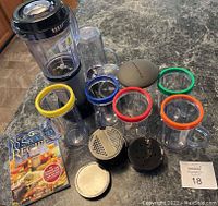 Magic Bullet blender base with multiple blending cups featuring colored silicone lip rings, various blade attachments, a flat storage lid, and a recipe guide booklet arranged on a countertop.